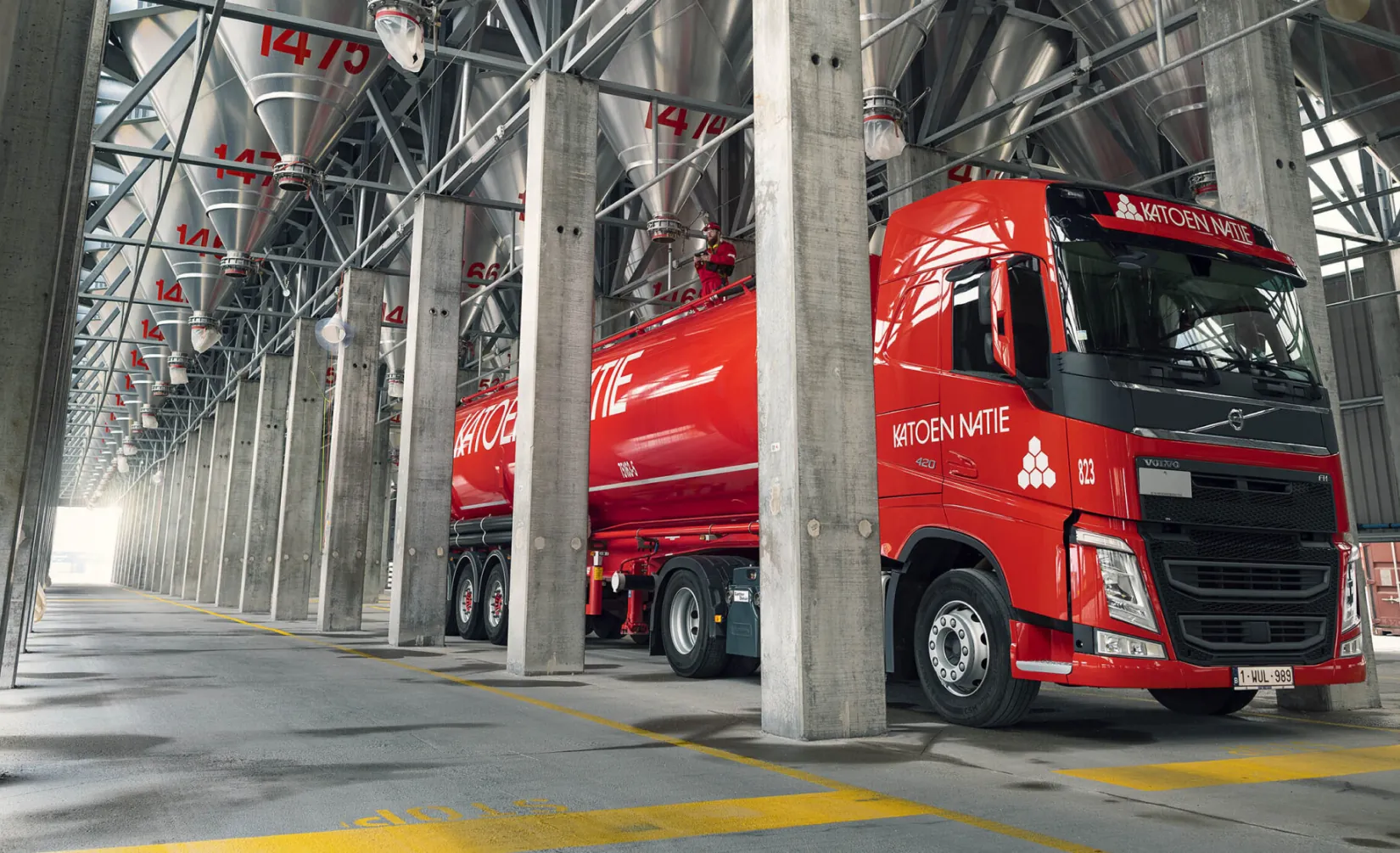 A red Katoen Natie tanker truck at an industrial loading facility